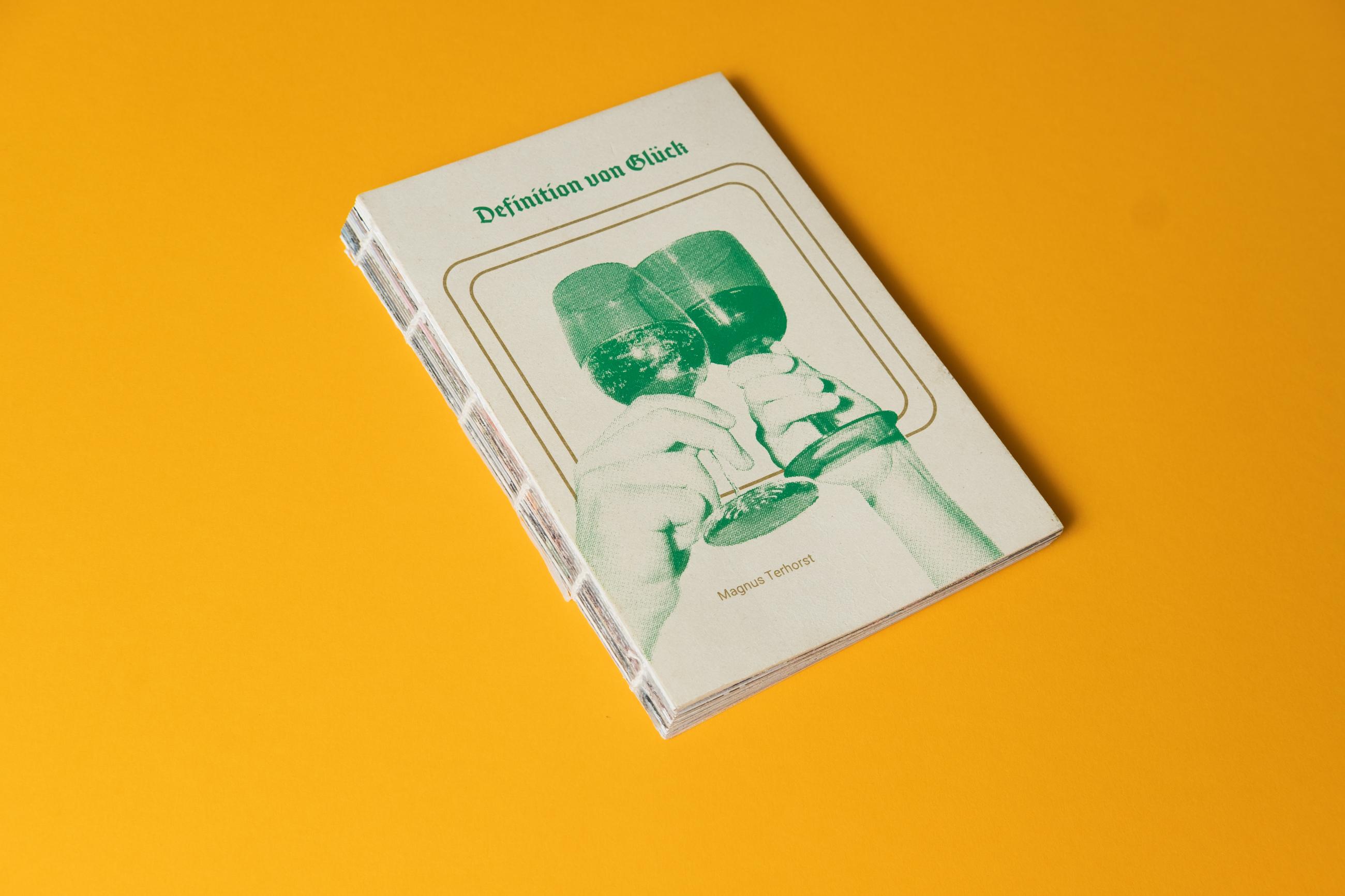 Das Foto zeigt das Cover des Buches vor gelben Hintergrund. Auf dem Cover sieht man zwei anstoßende Gläser in grün gesiebdruckt umrahmt von goldenen Outlines in Form eines quadratischen Bierdeckels. Darüber befindet sich der Titel des Buches Definition von Glück